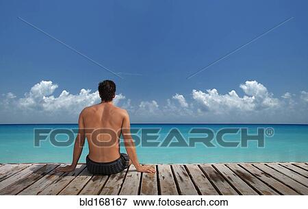 Stock Photo - Caucasian man sitting on wooden dock at ocean. Fotosearch