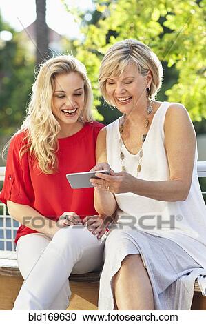 Stock Image - Caucasian mother and daughter using cell phone on bench. Fotosearch