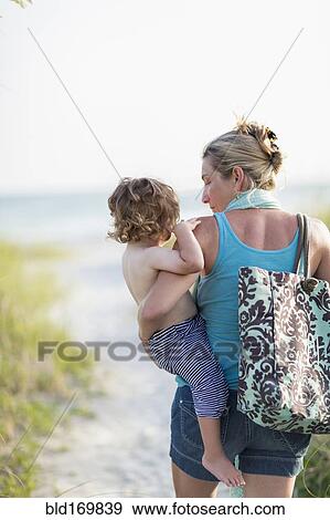 Stock Photo - Caucasian mother carrying baby son on beach. Fotosearch