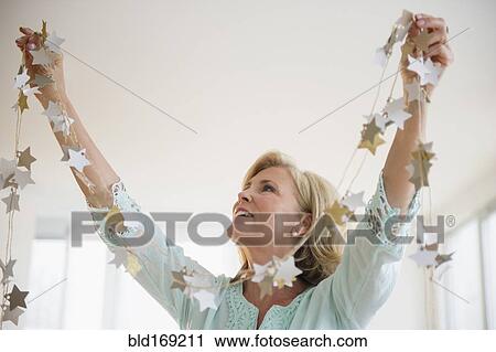Stock Image - Caucasian woman decorating with garland in living room. Fotosearch