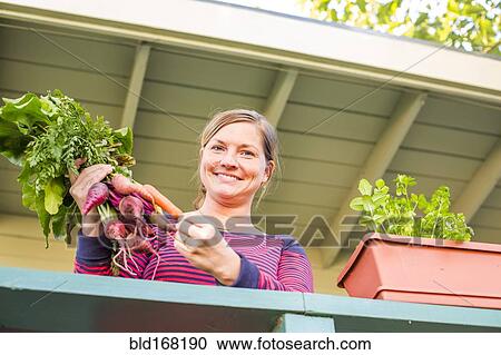 Stock Image - Caucasian woman holding fresh carrots on patio. Fotosearch
