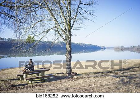 Stock Image - Caucasian woman sitting on bench near rural river. Fotosearch