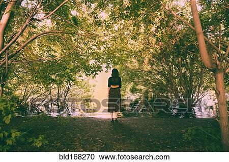Stock Image - Caucasian woman standing under trees near river. Fotosearch