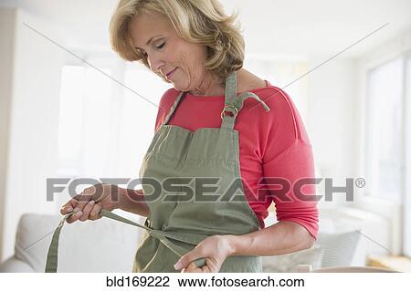 Stock Image - Caucasian woman tying apron in living room. Fotosearch