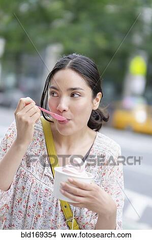 Picture - Hispanic woman eating frozen yogurt on city sidewalk. Fotosearch