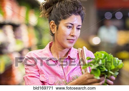 Stock Image - Hispanic woman examining produce at grocery store. Fotosearch