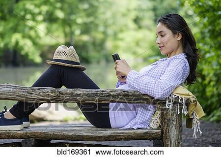 Stock Image - Hispanic woman using cell phone on park bench. Fotosearch