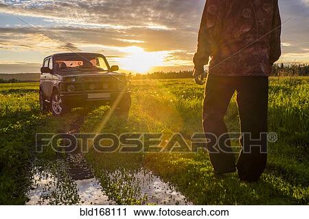 Stock Image - Mari man walking toward car at sunset in rural field. Fotosearch
