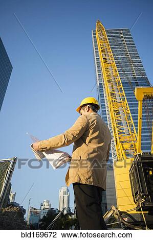 Stock Image - Mixed race architect reading blueprints at construction site. Fotosearch