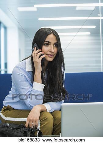 Stock Image - Mixed race businesswoman talking on cell phone in office lobby. Fotosearch