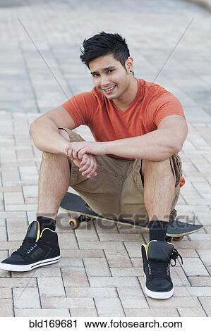 Stock Image - Mixed race man sitting on skateboard. Fotosearch