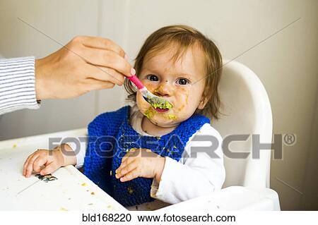 Stock Image - Mother feeding baby girl in high chair. Fotosearch