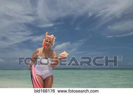 Stock Photograph - Older Caucasian woman holding conch shell on beach. Fotosearch