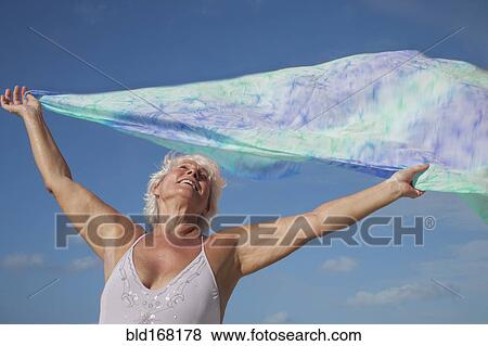 Stock Photo - Older Caucasian woman holding fabric in wind under blue sky. Fotosearch