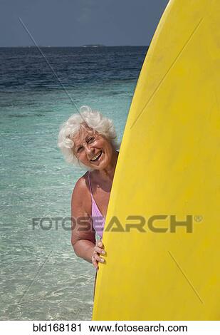 Stock Image - Older Caucasian woman holding surfboard on beach. Fotosearch