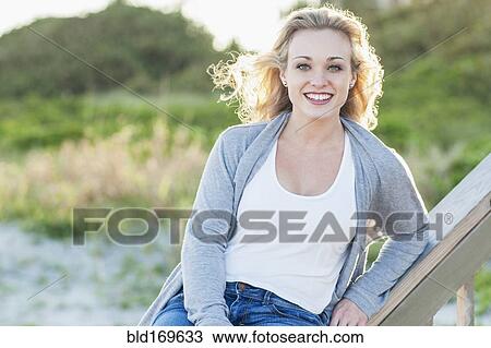 Stock Image - Smiling Caucasian woman sitting on banister over beach. Fotosearch
