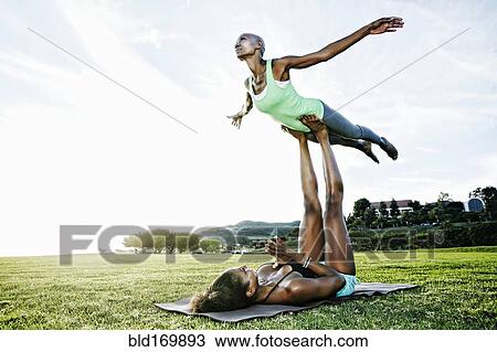 Stock Image - Woman and friend practicing acro yoga in park. Fotosearch