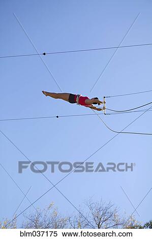 Stock Photography - Acrobat hanging from trapeze under blue sky. Fotosearch