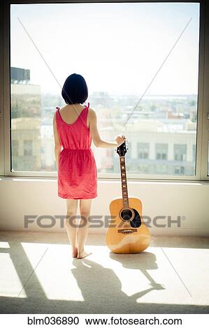 Stock Image - Asian woman holding guitar in window. Fotosearch