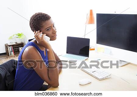 Stock Image - Black businesswoman talking on cell phone at office desk. Fotosearch