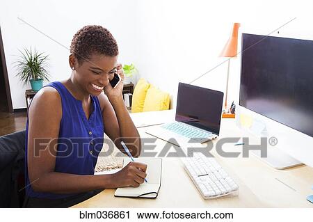 Stock Image - Black businesswoman talking on cell phone at office desk. Fotosearch