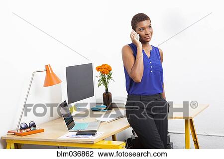 Stock Image - Black businesswoman talking on cell phone at office desk. Fotosearch