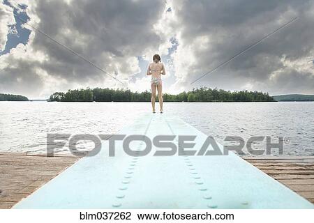 Stock Image - Caucasian girl standing on diving board over lake. Fotosearch