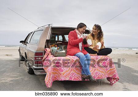 Stock Image - Caucasian mother and daughter eating picnic in car trunk on beach. Fotosearch