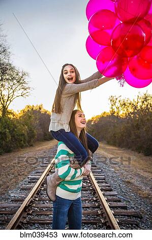 Stock Image - Caucasian teenage girl carrying sister on shoulders with balloons. Fotosearch