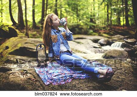 Picture - Caucasian teenage girl drinking coffee on forest rock. Fotosearch