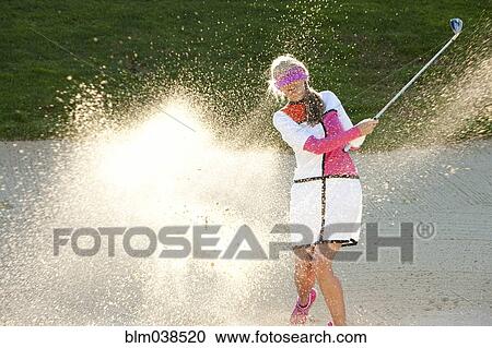 Stock Image - Caucasian woman hitting golf ball in sand trap while blindfolded. Fotosearch