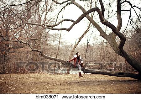 Stock Image - Caucasian woman sitting in tree in rural field. Fotosearch