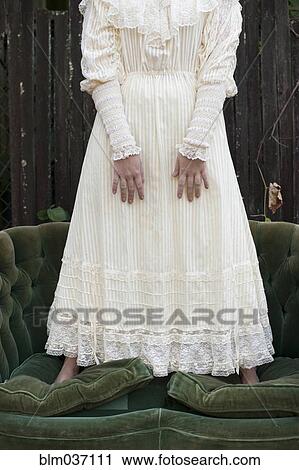 Stock Image - Caucasian woman wearing vintage gown standing on sofa. Fotosearch