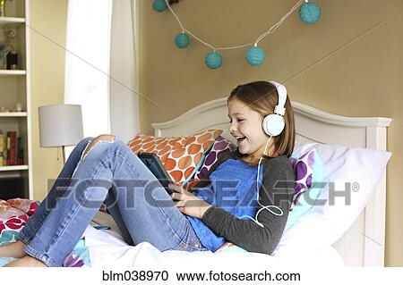 Stock Image - Girl listening to headphones with digital tablet on bed. Fotosearch