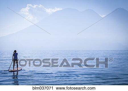 Stock Image - Girl standing on paddleboard in remote lake. Fotosearch