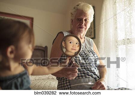 Stock Image - Grandmother holding mirror for granddaughter on bed. Fotosearch