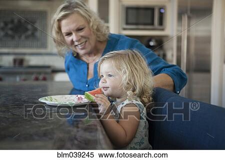Stock Photography - Grandmother watching granddaughter eating watermelon in kitchen. Fotosearch