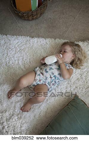 Stock Image - High angle view of preschooler girl drinking bottle on rug. Fotosearch