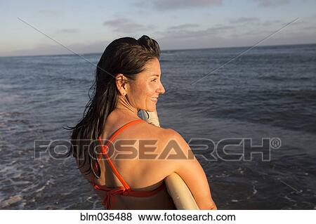 Stock Photo - Hispanic woman holding surfboard in ocean. Fotosearch