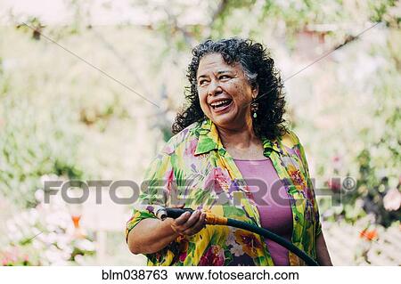 Stock Image - Hispanic woman watering plants in garden. Fotosearch