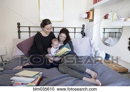 Stock Image - Lesbian couple reading to baby daughter on bed. Fotosearch