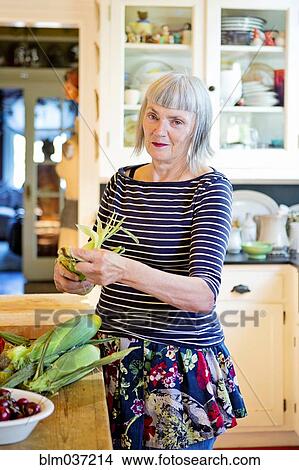 Picture - Older Caucasian woman shucking corn in kitchen. Fotosearch