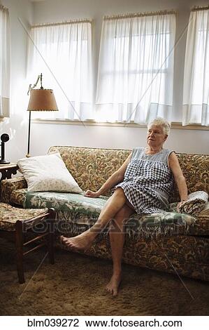 Stock Image - Older woman sitting on sofa in living room. Fotosearch