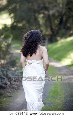 Stock Image - Rear view of bride walking on rural dirt road. Fotosearch