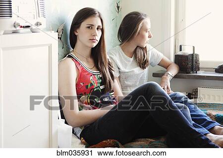 Stock Image - Serious sisters sitting on bed. Fotosearch