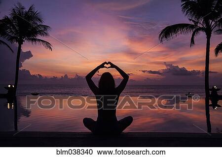 Stock Image - Silhouette of woman gesturing love near swimming pool at sunset. Fotosearch