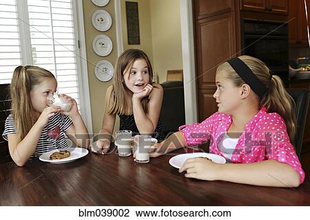 Stock Image - Sisters eating cookies with milk at table. Fotosearch