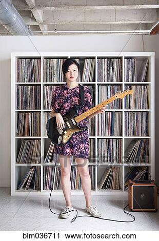 Stock Image - Taiwanese woman playing electric guitar near record collection. Fotosearch