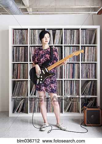 Stock Image - Taiwanese woman playing electric guitar near record collection. Fotosearch