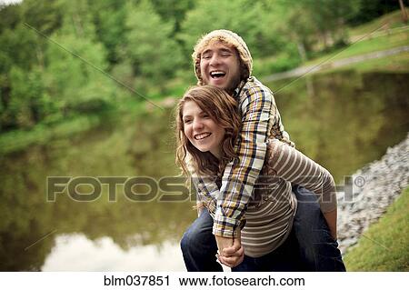 Stock Image - Woman carrying boyfriend piggyback near lake. Fotosearch